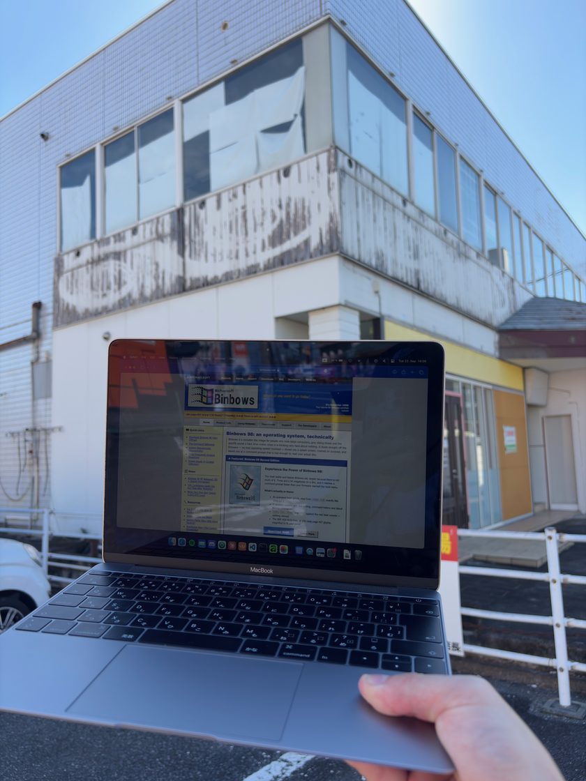 A hand holding a MacBook with binbows.net open in the browser, in front of the former shop building
