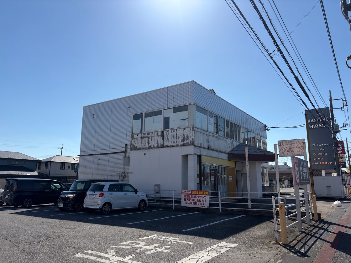 The same two-storey building in 2026, empty and without its sign. The weathered panel below the upper windows still shows faint outlines of the old lettering. Cars are parked in front under a clear blue sky.