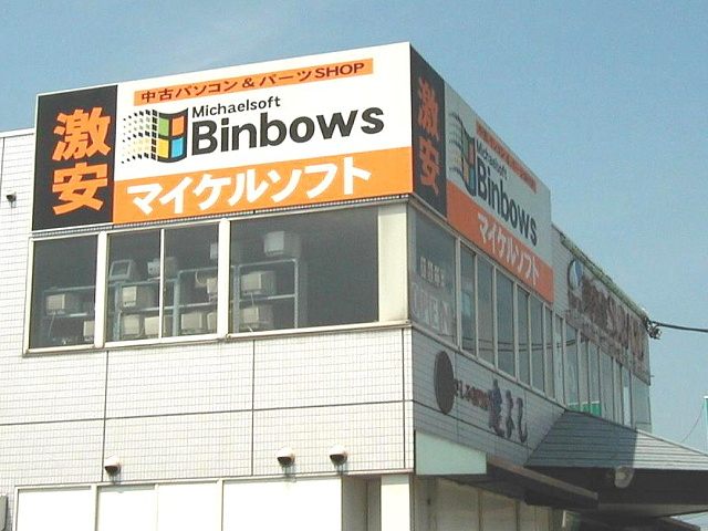 A Japanese used-computer shop with a large orange and black sign reading 'Michaelsoft Binbows' in the style of the Windows logo, with Japanese text マイケルソフト and 激安 meaning 'super cheap'